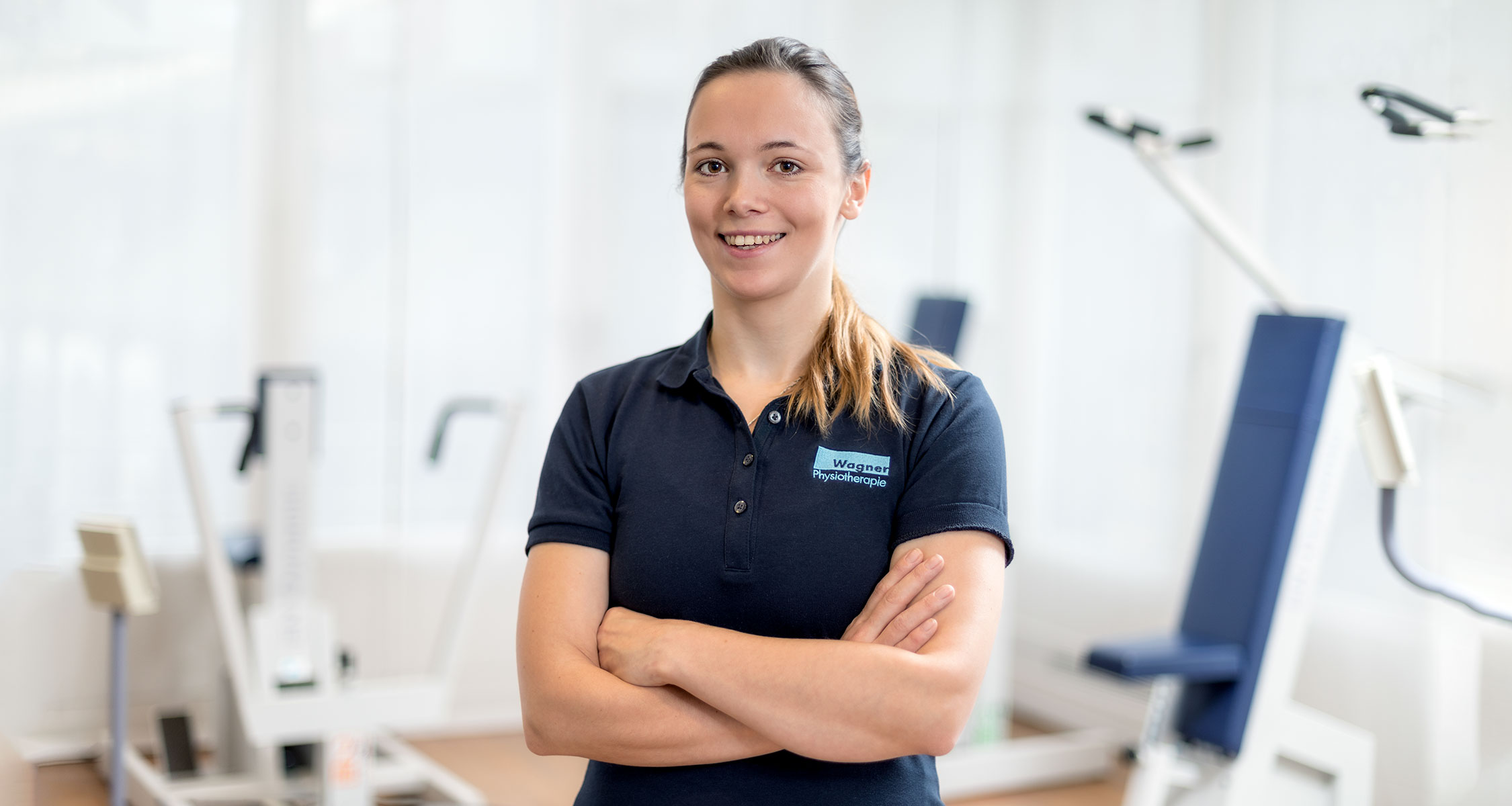 Freundliches Mitarbeiterportrait einer Physiotherapeutin mit verschränkten Armen in einer hellen Praxis.